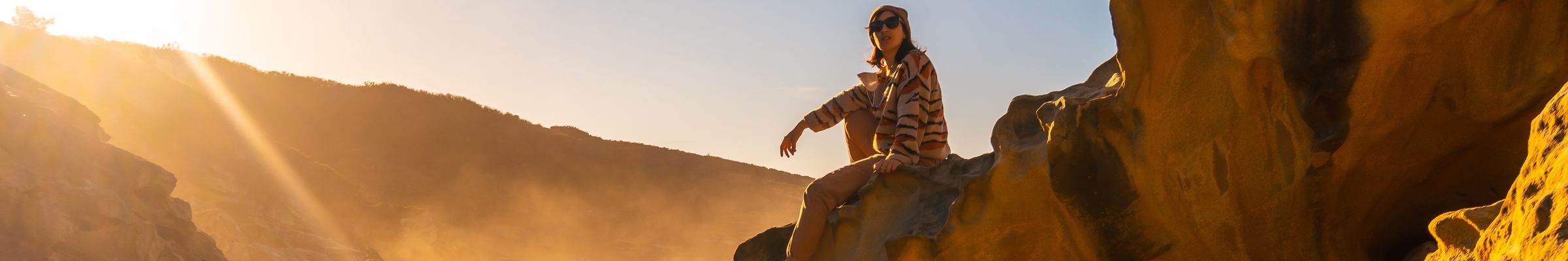 Two people on sunlit coastal rocks, one sitting on a ledge above the sea